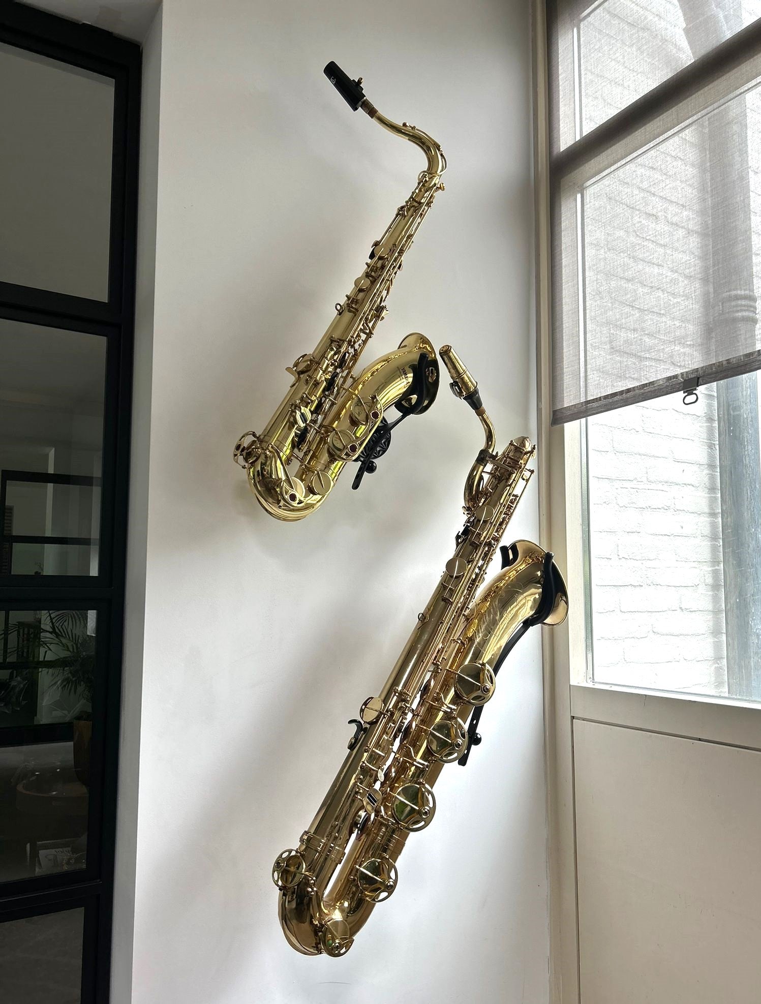 Two gold saxophones mounted on a white wall with a window in the background.