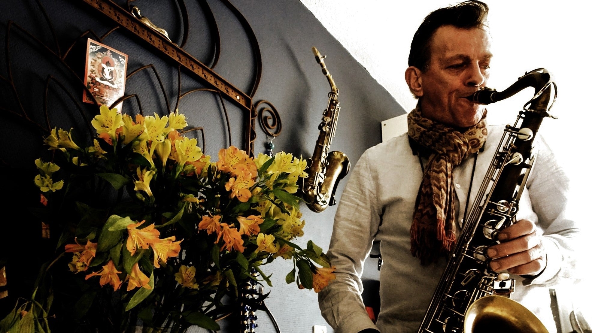 Man playing saxophone at home beside flowers and wallmounted stand with alto saxophone.